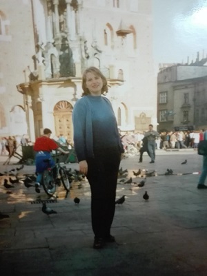 Dziewczyna w niebieskim sweterku i czarnych spodniach pozuje do zdjęcia na krakowskim rynku, pod wieżą Kościoła Mariackiego