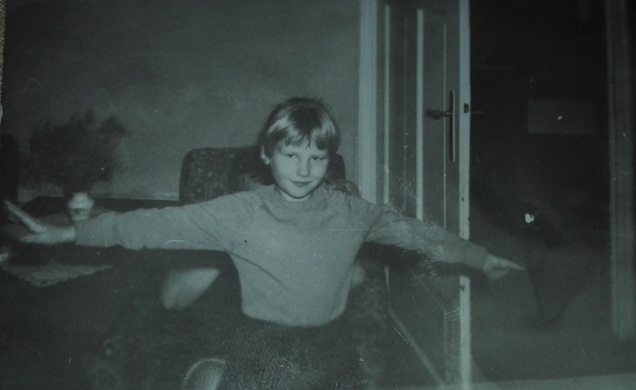 Stare, czarno - białe zdjęcie dziewczynki o rozłożonych ramionach i delikatnym uśmiechu, na tle fragmentu prywatnego mieszkania