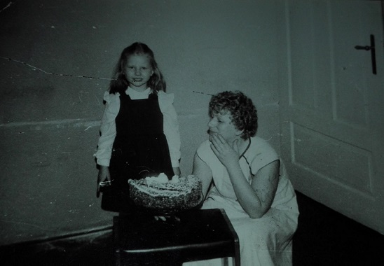 Stare czarno białe zdjęcie przedstawia dziewczynkę w czarnej sukience pozującą obok swojej mamy i urodzinowego tortu.