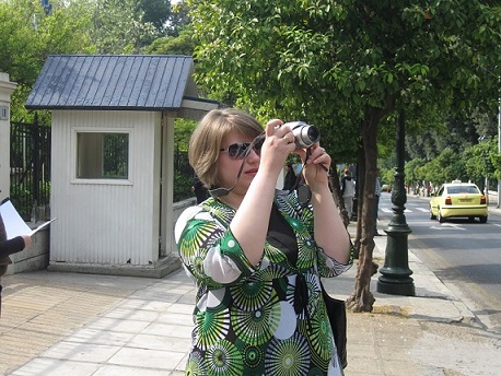 Młoda kobieta w okularach przeciwsłonecznych wykonuje zdjęcie aparatem cyfrowym, w tle porośnięta drzewami ulica.