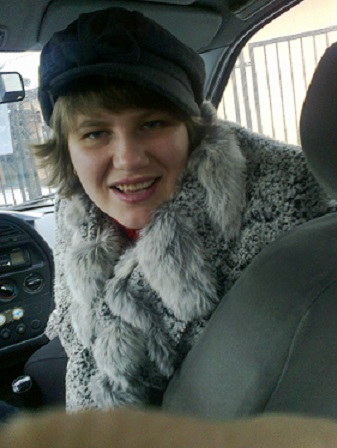 Młoda, usmiechnięta kobieta w zimowej czapce "na bakier" i zimowym płaszczu pozuje w półobrocie na przednim siedzeniu pasażera samochodu. Zdjęcie wykonane przez pasażera z tylnego siedzenia.